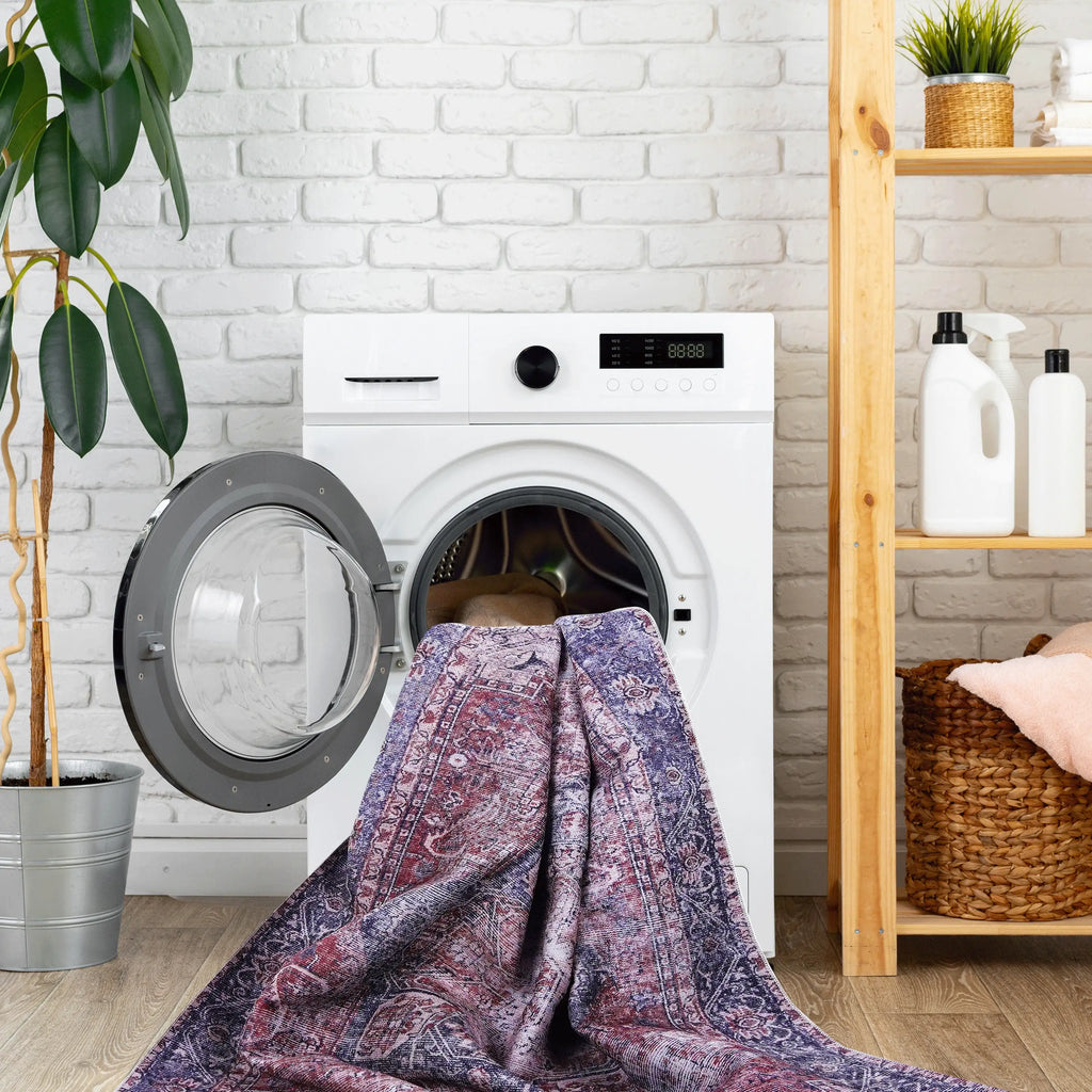 A white front-loading washing machine with its door open, shown loading a patterned purple and blue blanket or towel. The appliance is set in a bright laundry room with a white brick wall, a potted plant, and a wooden shelf with cleaning supplies.