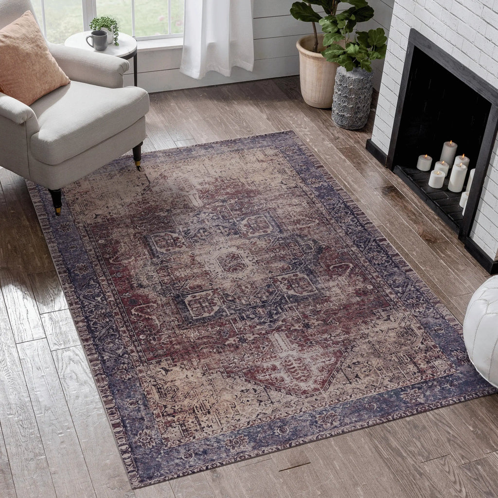 A distressed vintage-style area rug with a faded medallion pattern in muted purple, beige, and brown tones, styled on a light wood floor in a bright living room next to a white armchair and fireplace.