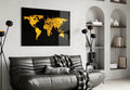 A rectangular black wall art print featuring a glowing gold world map, displayed above a dark gray leather sofa in a modern living room setting.