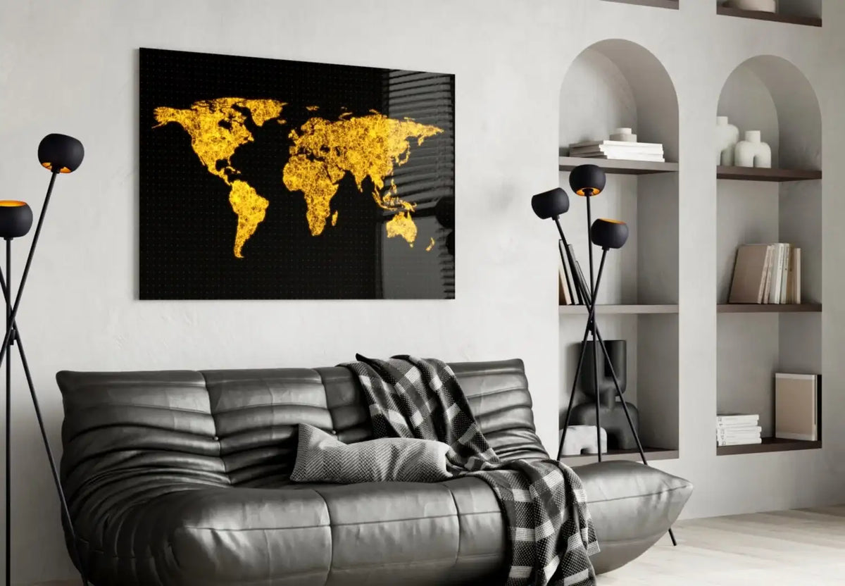 A rectangular black wall art print featuring a glowing gold world map, displayed above a dark gray leather sofa in a modern living room setting.
