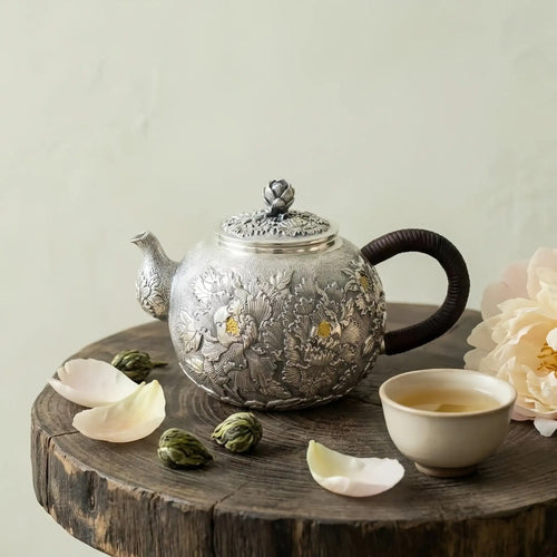 Decorative silver teapot with floral patterns on a wooden surface with tea leaves and a cup.